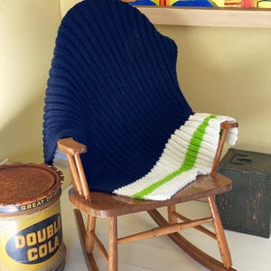 May include: A navy blue knitted throw blanket with white and green stripes draped over a wooden rocking chair. The chair is in a room with a yellow wall and a painting on the wall behind it.