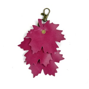 May include: A burgundy leather keychain featuring a cluster of three leaf-shaped cutouts. The leaves are layered and attached to a bronze-colored clasp and a small rivet. The keychain is designed to be a decorative accessory.