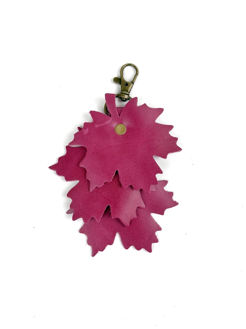 May include: A burgundy leather keychain featuring a cluster of three leaf-shaped cutouts. The leaves are layered and attached to a bronze-colored clasp and a small rivet. The keychain is designed to be a decorative accessory.
