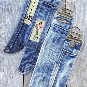 Puede incluir: Tres llaveros de mezclilla hechos de jeans azules reciclados. Cada correa cuenta con un cierre de metal con anillo, un botón y bordes deshilachados. Una correa muestra un parche con el texto 20X STYLE 01 COMPETITION y otra tiene una etiqueta Wrangler 20X.