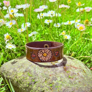 May include: A brown leather cuff bracelet with a floral design, including a sunflower and ladybug. The bracelet is set on a mossy rock, surrounded by daisies and green grass.