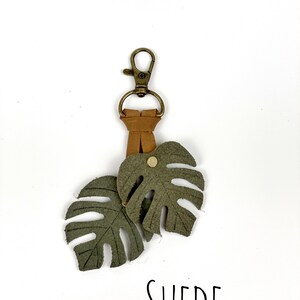 May include: A green suede keychain with two monstera leaf charms and a gold metal key ring. The keychain has the word "SUEDE" printed on the white background.