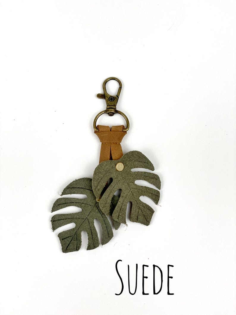 May include: A green suede keychain with two monstera leaf charms and a gold metal key ring. The keychain has the word "SUEDE" printed on the white background.