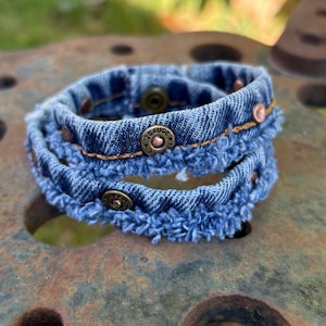 May include: A wrap bracelet crafted from blue denim fabric with frayed edges. The design features metallic 12 Gauge shotgun shell casing accents and bronze-toned snap button closures, creating a layered appearance with visible orange stitching.