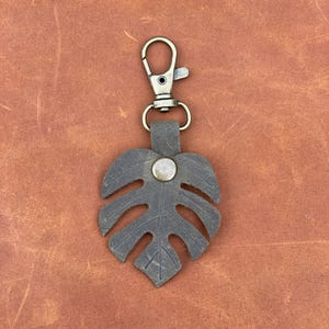 May include: A dark brown leather keychain with a monstera leaf design and a silver metal button. The keychain has a brass-colored metal clasp.