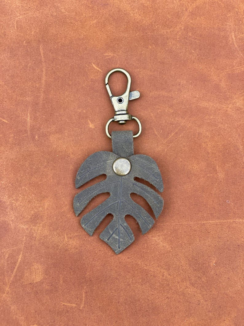 May include: A dark brown leather keychain with a monstera leaf design and a silver metal button. The keychain has a brass-colored metal clasp.