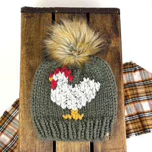 May include: A chunky knit olive green beanie featuring a white rooster graphic with a red comb and yellow feet. The hat is topped with a large, fluffy, multi-toned brown and tan faux fur pom-pom and includes a small brown tag on the ribbed brim.