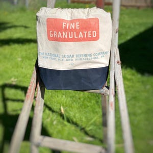 May include: A white canvas tote bag with a red and blue label that reads "FINE GRANULATED THE NATIONAL SUGAR REFINING COMPANY NEW YORK, N.Y. AND PHILADELPHIA, PA."