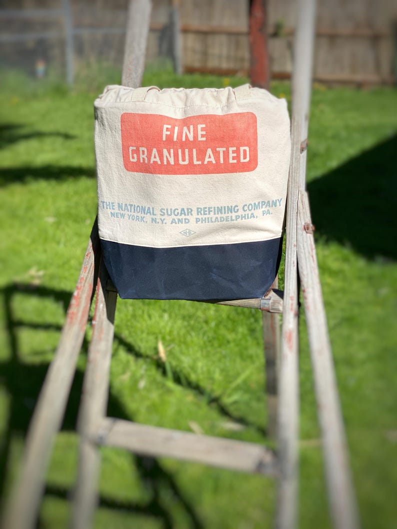 May include: A white canvas tote bag with a red and blue label that reads "FINE GRANULATED THE NATIONAL SUGAR REFINING COMPANY NEW YORK, N.Y. AND PHILADELPHIA, PA."
