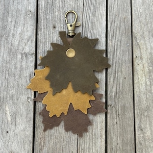 May include: A leather keychain featuring three maple leaf cutouts in shades of brown and tan. The leaves are layered and attached to a brass-colored clasp. The keychain is displayed on a weathered wooden surface.