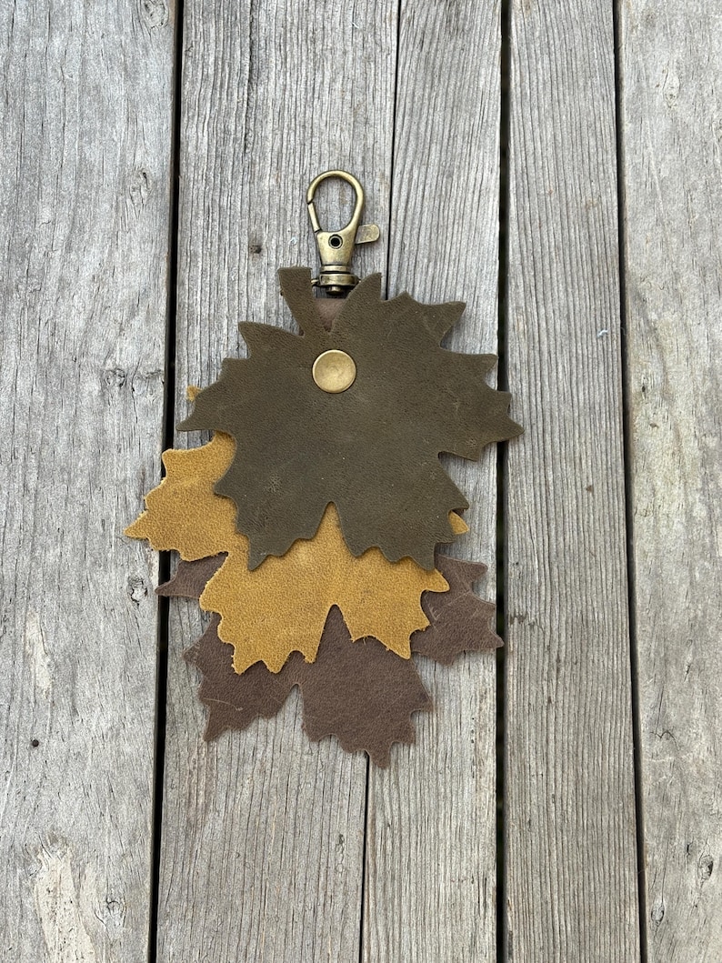 May include: A leather keychain featuring three maple leaf cutouts in shades of brown and tan. The leaves are layered and attached to a brass-colored clasp. The keychain is displayed on a weathered wooden surface.