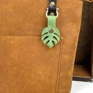 May include: A brown leather tote bag with a green leather monstera leaf keychain. The bag has a zipper closure and a brown leather strap.
