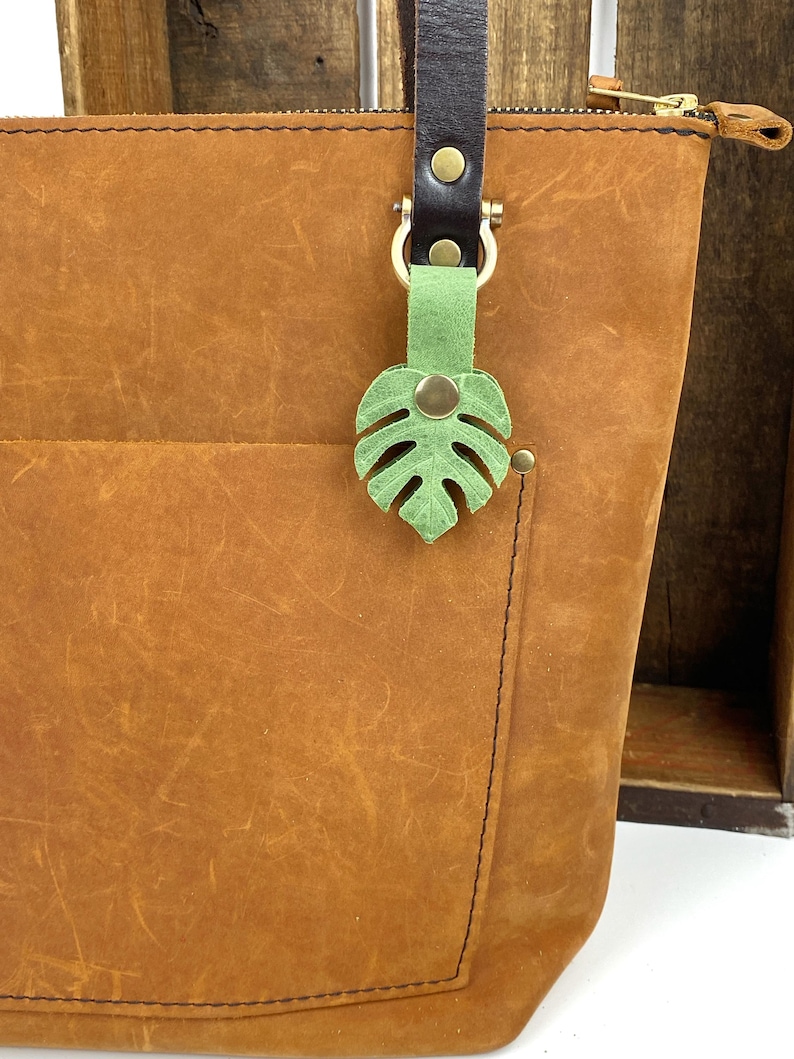 May include: A brown leather tote bag with a green leather monstera leaf keychain. The bag has a zipper closure and a brown leather strap.