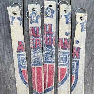 May include: Five rectangular keychains with metal rings. The keychains are made of a beige fabric with a distressed "All American" design in red and blue. The keychains are arranged vertically on a wooden surface.