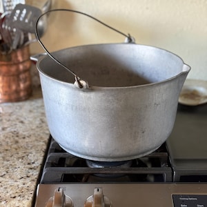 May include: A silver-colored, vintage cooking pot with a black handle sits on a gas stove. The pot has a rounded shape and two small side handles. The stove controls are visible below the pot.