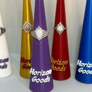 May include: Close-up of several rings on display. The rings are silver-toned with various gemstone designs. They are displayed on colorful, cone-shaped stands. The stands are white, gold, purple, red, and blue, each with the text "Horizon Goods".