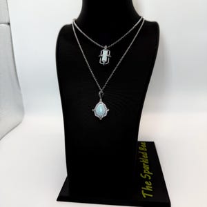 May include: Two silver necklaces displayed on a black mannequin. The top necklace has a small, rectangular pendant. The bottom necklace features a teardrop-shaped, light blue pendant with a halo of small, clear stones. The necklaces are on a black stand.