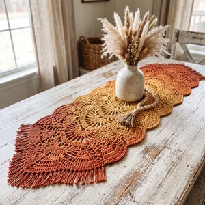 May include: A crochet table runner featuring an ombre color gradient transitioning from deep orange to yellow. The runner has a scalloped edge with fringe at both ends. A speckled ceramic vase filled with dried grasses sits on the runner.