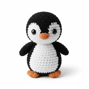 May include: A handmade crochet penguin toy. The penguin has a black head and wings, a white belly, and orange feet and beak. The toy has large, black safety eyes. The penguin is standing upright on a white background.