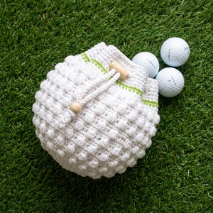 May include: A white crocheted drawstring pouch featuring a textured bobble stitch pattern and a thin lime green horizontal stripe. The pouch includes a white braided cord with a light wooden bead closure and three white golf balls nearby.
