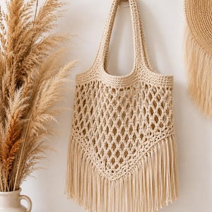 May include: A beige crochet tote bag featuring an open diamond mesh pattern and a V-shaped bottom edge finished with long, straight fringe. The bag has two integrated shoulder straps and is constructed from a light-coloured cord material.