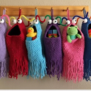 以下が含まれることがあります： 木製のラックに吊るされた、カラフルな編み物で作られたモンスター型の収納バッグが6つ。各バッグには、動く目、モールで作られた触角、フリンジが付いています。バッグの中には、アヒルのオモチャ、ブロック、カエル、リングスタッカーなどのおもちゃが入っています。