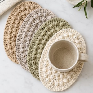 May include: A set of four oval-shaped crochet coasters in tan, light gray, sage green, and cream, each featuring a textured bobble stitch pattern. A speckled cream ceramic mug sits on top of the cream-colored coaster, showcasing the layered design.