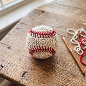 May include: A spherical baseball crafted from off-white yarn with prominent red stitched detailing. The ball features a textured crochet pattern throughout its surface, mimicking the appearance of a traditional baseball with its signature red lacing.