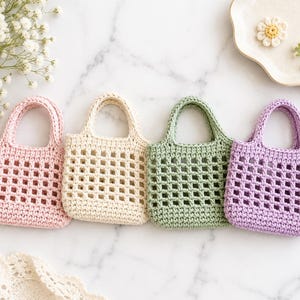 May include: Four small, square-shaped crochet bags arranged in a row. Each bag features a solid base and a grid-like openwork pattern on the body, finished with two short handles. The bags are colored in light pink, cream, sage green, and lavender.