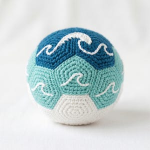 May include: A crocheted ball with a wave design. The ball is made of blue, teal, and white yarn. White yarn is used to create wave patterns on the surface of the ball. The ball is a sphere shape.