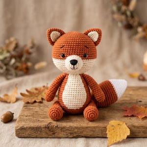 May include: A crocheted fox toy featuring a burnt orange body, a cream-coloured chest and muzzle, and a bushy tail with a white tip. The fox has small black eyes, a stitched black nose, and pointed ears with cream-coloured inner details.