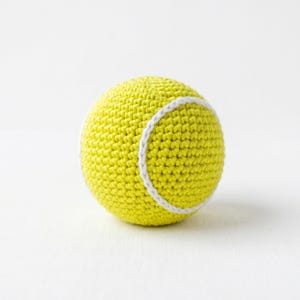 Könnte beinhalten: Ein kugelförmiger Tennisball, der aus leuchtend gelbem Garn gehäkelt wurde. Der Ball weist die charakteristische, geschwungene weiße Garnnaht auf, die um die Mitte gestickt ist und eine strukturierte Oberfläche mit einem dichten Maschenbild erzeugt.