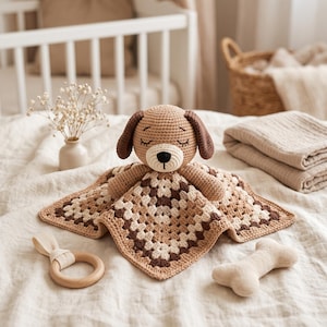 May include: A crochet security blanket featuring a stuffed dog head with closed eyes and brown ears, attached to a square granny square blanket in shades of beige, cream, and dark brown. The toy is accompanied by a wooden ring and a bone-shaped toy.