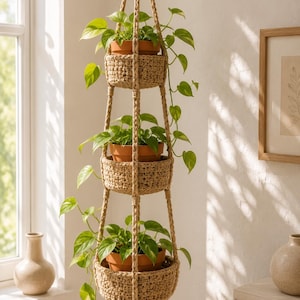 May include: A three-tiered hanging planter made of beige woven cord, featuring three circular baskets that hold terracotta pots with green trailing pothos plants. The structure is suspended by braided ropes and finished with a tassel at the bottom.