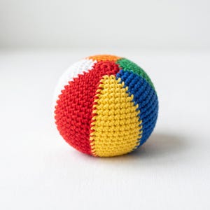May include: A spherical ball crafted from crocheted yarn featuring vertical segments in vibrant colors including red, yellow, blue, green, and white. The texture is consistent throughout the surface, showing the individual stitches of the yarn.