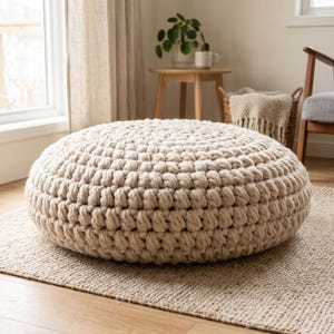 May include: A round, floor-based ottoman featuring a thick, chunky knit construction in a neutral beige color. The piece is crafted with a consistent, repeating loop pattern that creates a textured, soft surface across the entire exterior.
