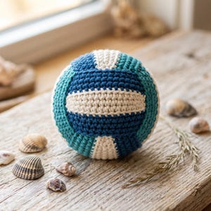 May include: A spherical crochet ball featuring a color-blocked pattern of dark blue, teal, and cream yarn. The textured surface is created with tight, uniform stitches, forming a classic volleyball-inspired design with alternating panels.
