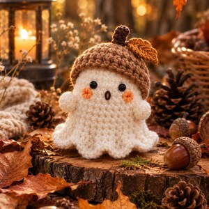 May include: A small, cream-colored crocheted ghost figure wearing a brown textured acorn cap on its head. The ghost features two black bead eyes, a small embroidered mouth, and two orange stitched circles on its cheeks, with a single brown leaf.