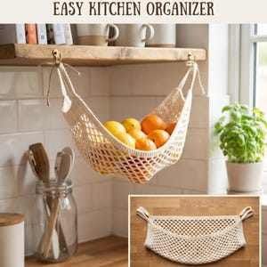 Puede incluir: Una hamaca para frutas tejida a ganchillo en color crema con un patrón de malla de rombos, suspendida con cuerda de dos ganchos de latón bajo un estante de madera, conteniendo naranjas y limones. Texto: HANGING FRUIT HAMMOCK.