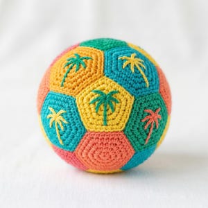 May include: A colorful, crocheted ball with a soccer ball design. The ball features a patchwork of hexagonal and pentagonal panels in various colors, including yellow, green, blue, orange, and pink. Each panel has a palm tree embroidered on it.