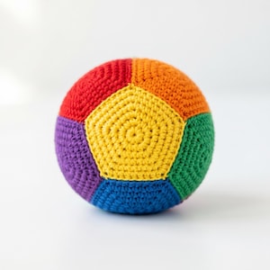 May include: A spherical toy ball constructed from crocheted yarn in a vibrant rainbow colour palette. The surface features a geometric pattern of pentagonal and hexagonal panels in red, orange, yellow, green, blue, and purple, creating a textured finish.