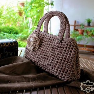 Puede incluir: Bolso de mano de ganchillo marrón hecho a mano con un adorno floral. El bolso tiene una forma estructurada con asas resistentes y una flor decorativa en la parte delantera. El bolso está sobre una tela marrón en una mesa de madera.