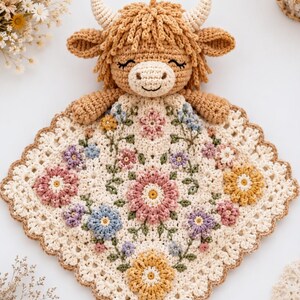 May include: A crochet security blanket featuring a tan highland cow head with small horns and closed eyes attached to a square cream-colored base. The blanket is decorated with a floral pattern of pink, yellow, blue, and purple crocheted flowers.