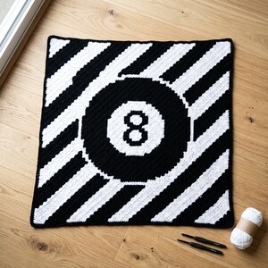 May include: A square crochet blanket featuring a black and white diagonal striped pattern with a large central black circle containing a white number eight. Two black crochet hooks and a ball of white yarn are positioned near the blanket.