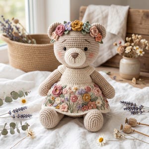 May include: A beige crochet teddy bear featuring a floral crown made of small yarn flowers in shades of pink, yellow, and blue. The bear wears a cream-coloured crochet dress adorned with an intricate, colourful embroidered floral pattern.