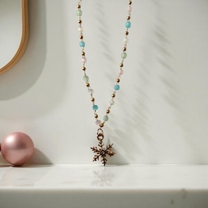 Puede incluir: Un delicado collar con una cadena dorada y cuentas pastel en rosa, azul, verde y blanco. Un colgante de copo de nieve cuelga de la cadena. El collar se exhibe sobre una superficie blanca.