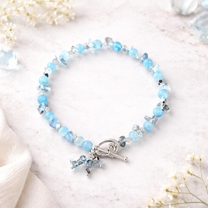 May include: A bracelet with light blue and clear beads, with silver-toned accents. The bracelet has a toggle clasp and a small cluster of blue beads as a charm. The jewellery is displayed on a white surface.