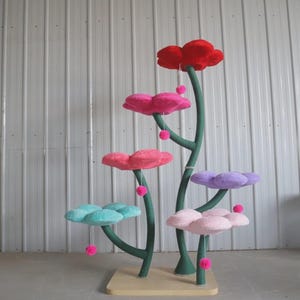 Könnte beinhalten: Ein skurriler Katzenbaum mit floralem Design. Der Baum hat mehrere wolkenförmige Plattformen in Rot-, Rosa-, Lila- und Türkistönen, die von grünen Stielen getragen werden. Jede Plattform hat einen hängenden rosa Pompon.
