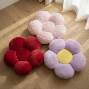 Könnte beinhalten: Drei plüschige, blumenförmige Kissen mit weicher, flauschiger Textur. Ein Kissen ist hellrosa, eines dunkelrot und eines hellviolett mit einem leuchtend gelben Zentrum. Jedes Kissen hat fünf abgerundete Blütenblätter um einen runden Kern.