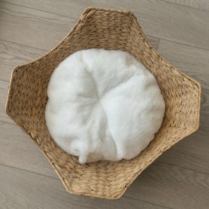 Könnte beinhalten: Ein sechseckiger geflochtener Korb aus hellbraunen Naturfasern mit einem weichen, flauschigen, runden Kissen aus weißem Kunstfell im Inneren. Der Korb hat eine strukturierte, geflochtene Oberfläche und ein offenes Design.
